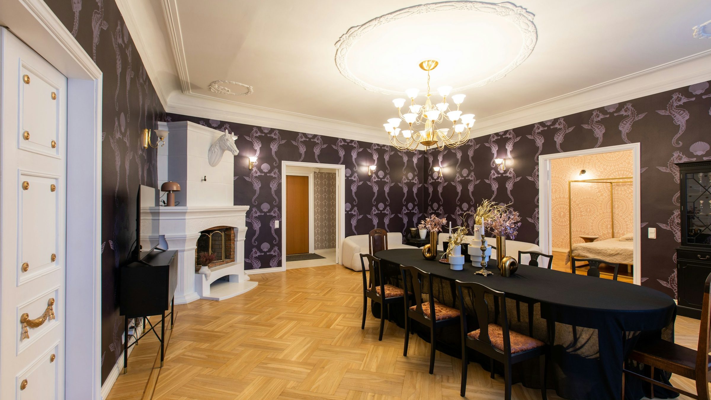 Dining room with deep aubergine seahorse-motif wallpaper, a white fireplace, brass chandelier and a long black dining table