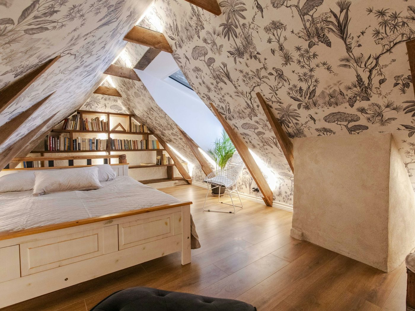 Attic bedroom with grey toile wallpaper on the gabled walls, exposed timber beams, a white wooden bed and a built-in bookshelf under the eaves