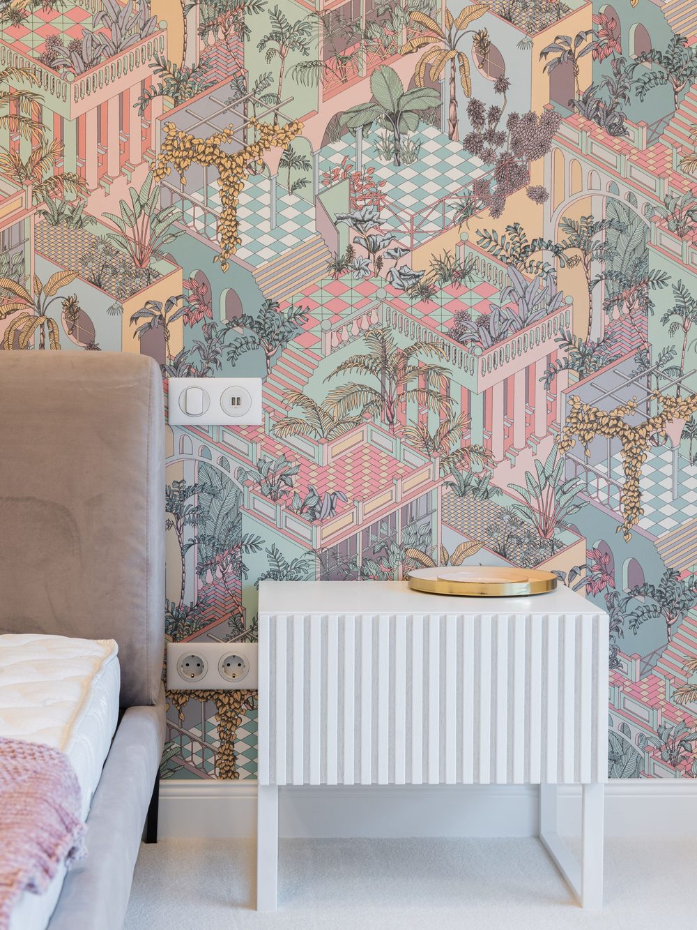 Bedroom corner with a pastel illustrated jungle-and-architecture wallpaper, a white fluted nightstand and the edge of an upholstered headboard
