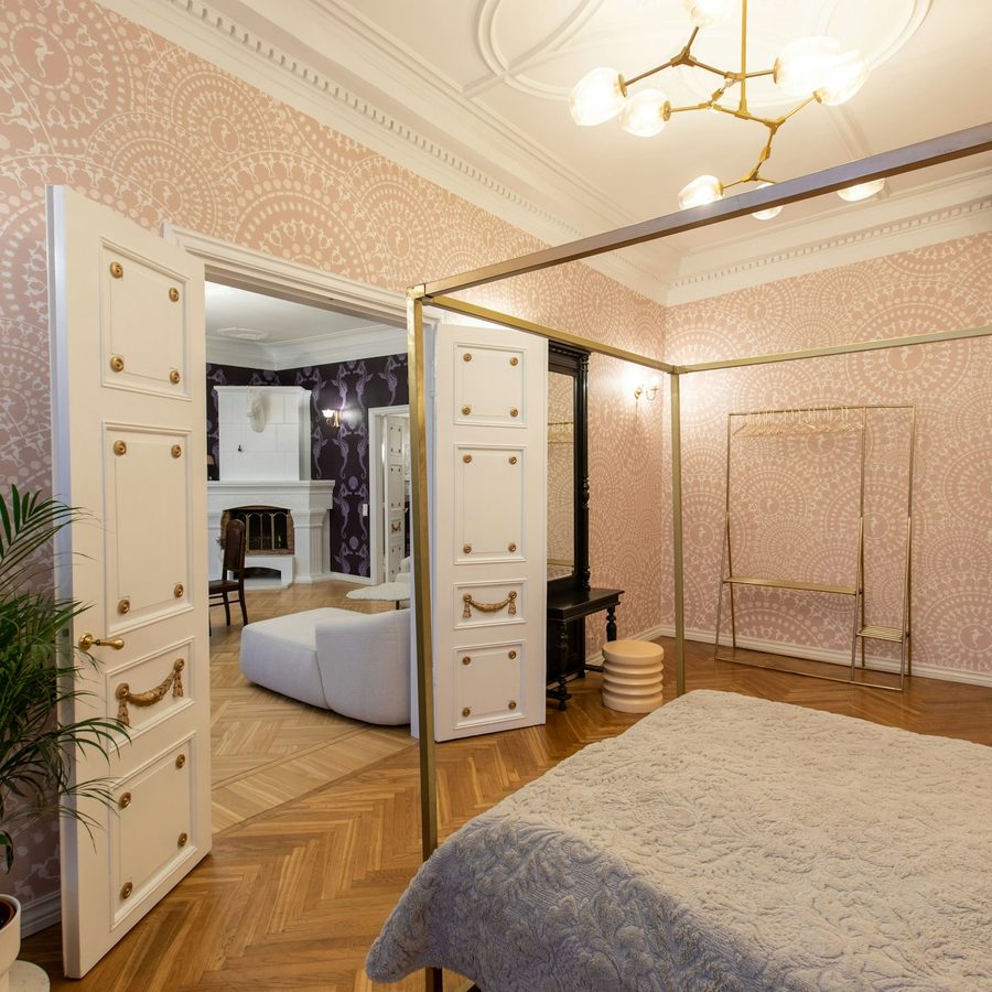 Bedroom with blush pink lace-medallion wallpaper, white double doors and a gold four-poster bed frame
