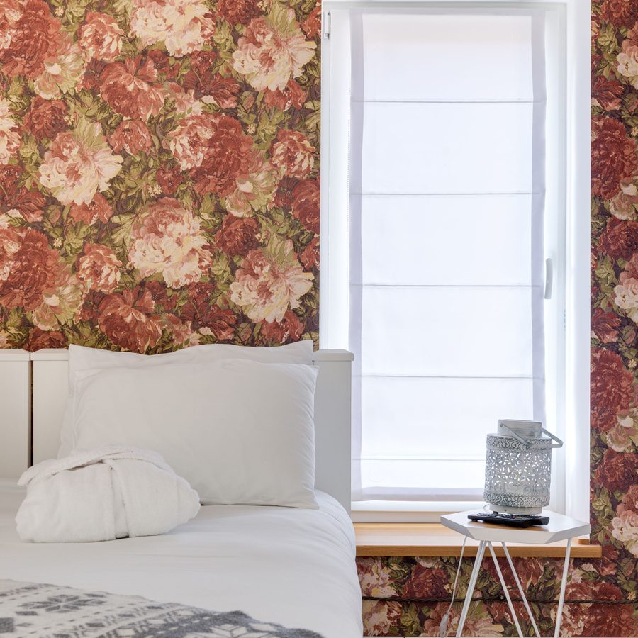 Bedroom wall covered in a bold red and pink rose wallpaper around a window with a white roman shade