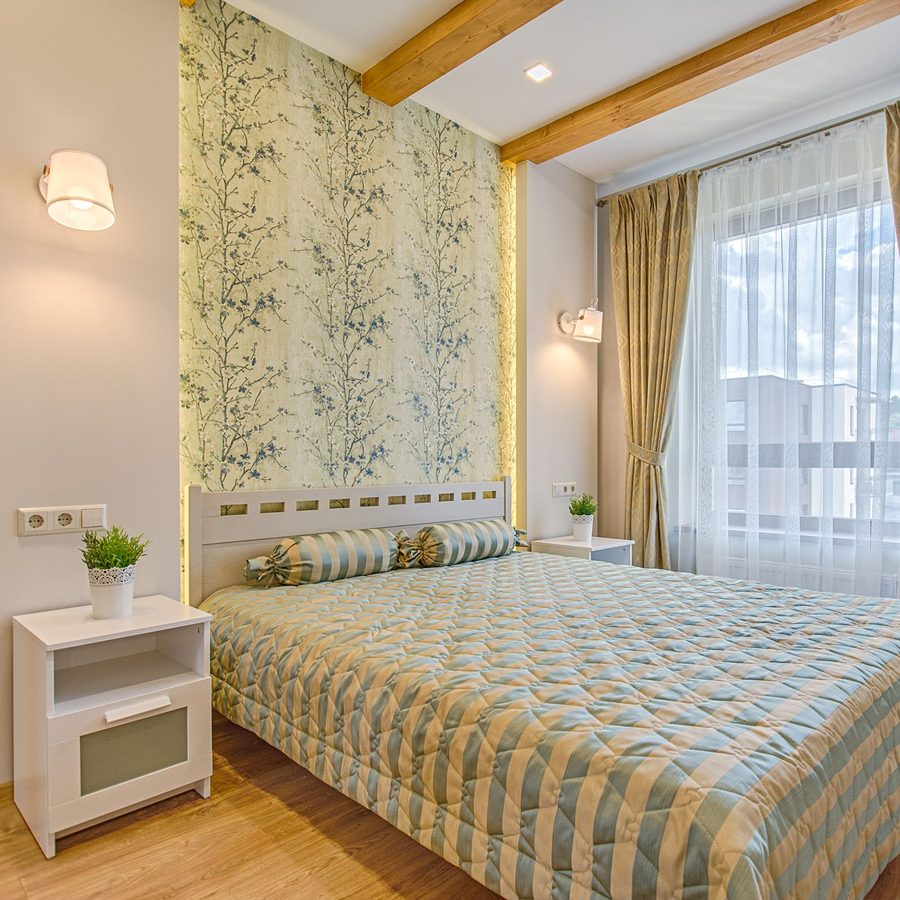 Bright bedroom with a sage botanical wallpaper panel behind a white bed, gold curtains and timber ceiling beams